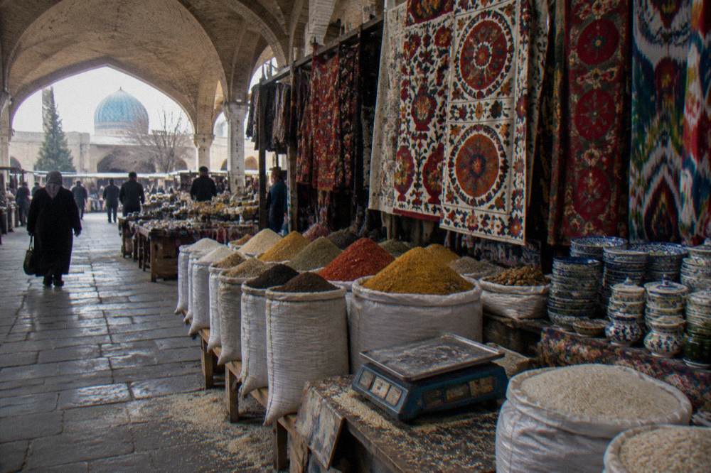 Covered market hall with hanging textiles, spices in sacks and stacked ceramics under vaulted brick ceilings