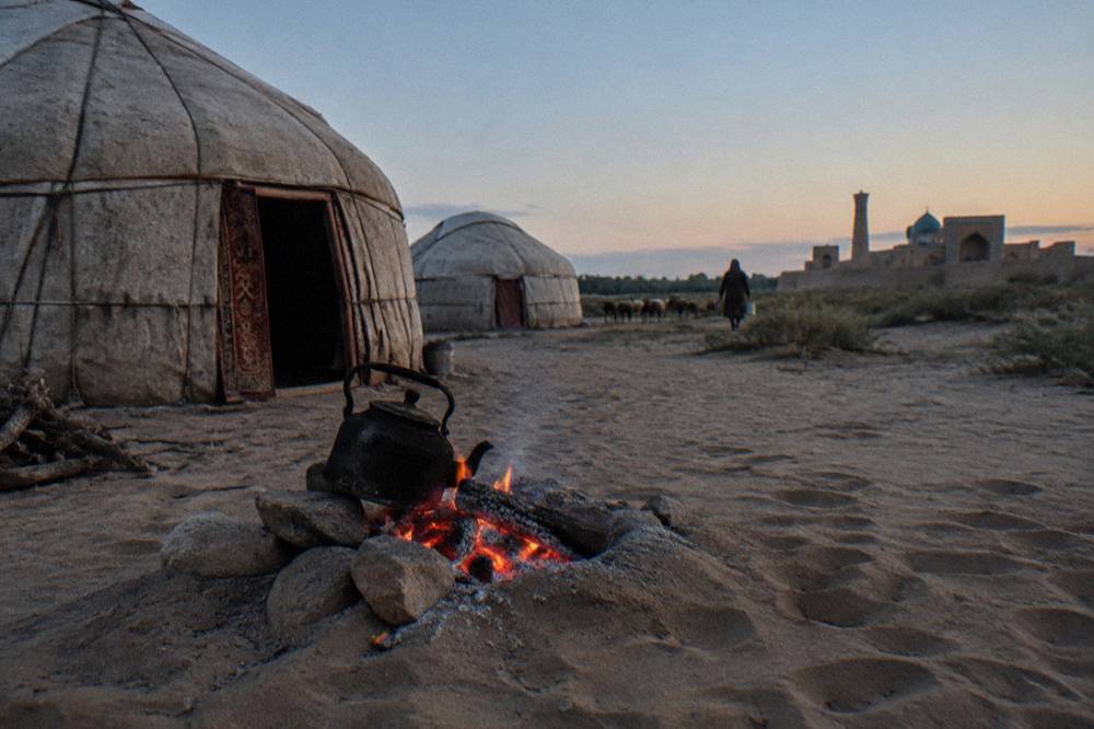 A campfire burning beside felt yurts on open sandy ground as evening falls