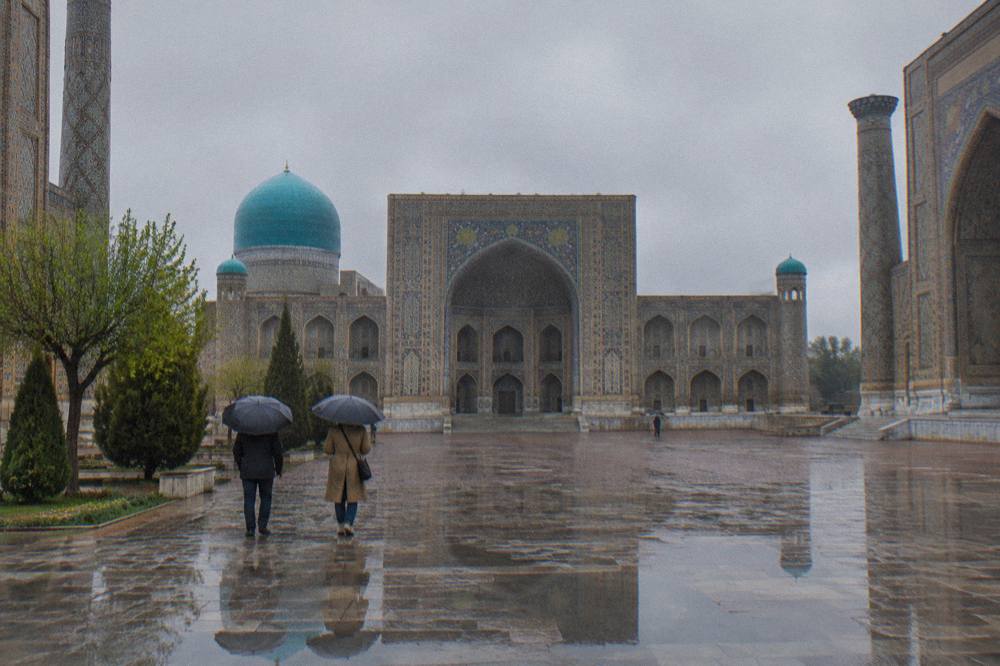 Travelers walking on a wide plaza beside a tiled madrasah facade in daylight