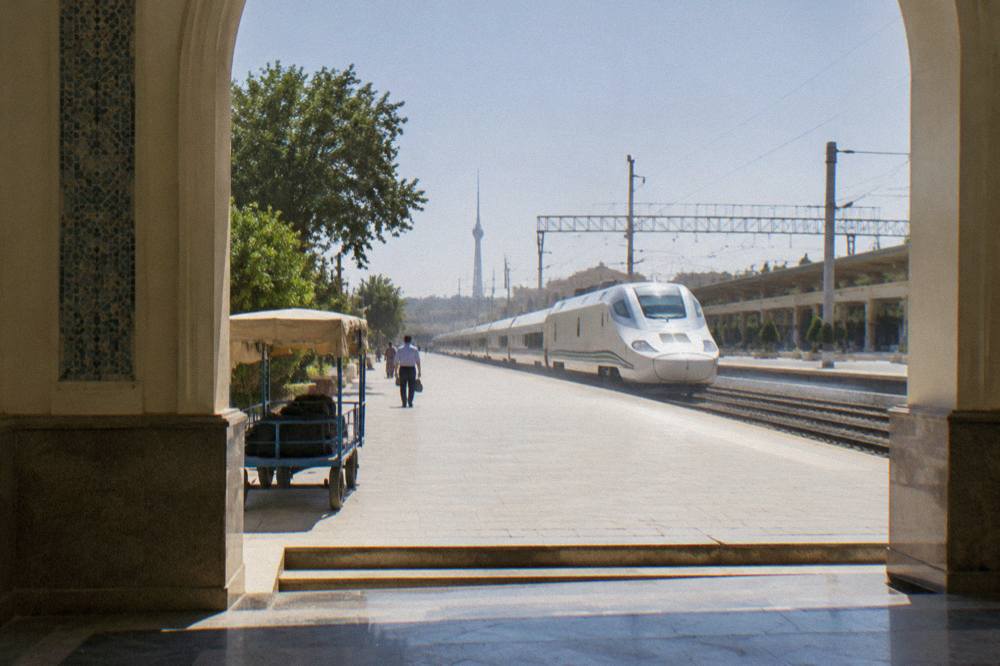 A modern white high-speed train waiting at a sunny platform beside a station building in Uzbekistan