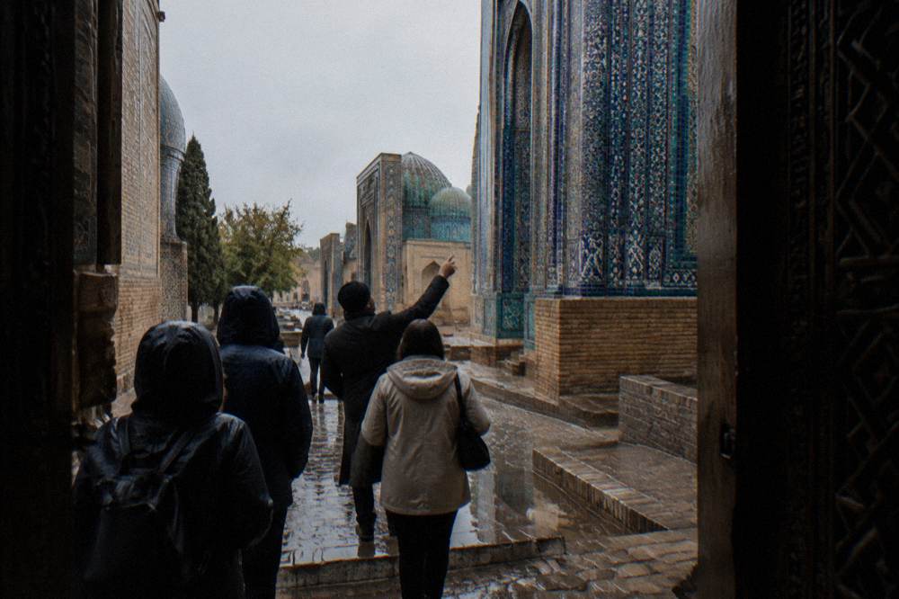 Guide speaking to visitors beside tiled medresa walls in a historic square